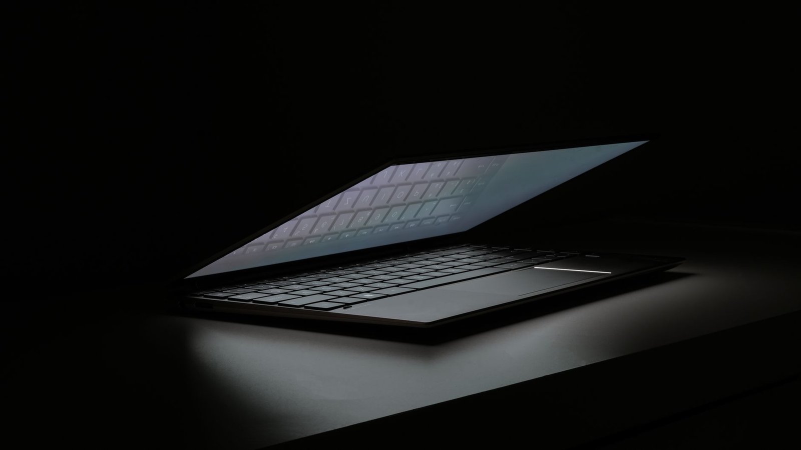Partial view of a laptop in a dimly lit room, showcasing its sleek design and illuminated keyboard.